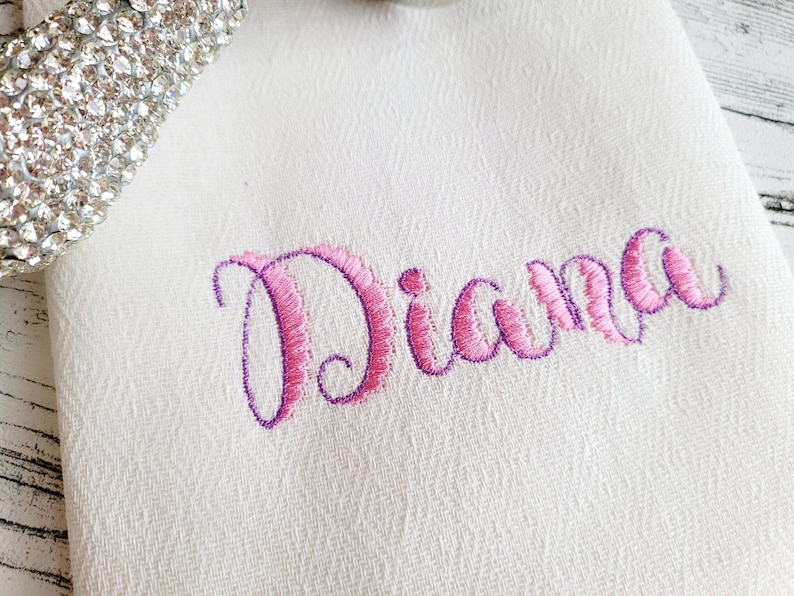 May include: White fabric with the name "Diana" embroidered in pink and purple cursive lettering.