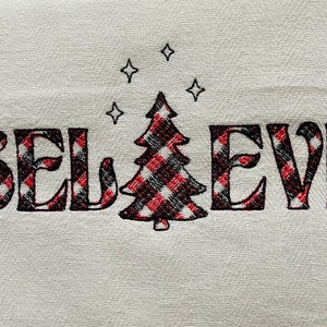 May include: A white kitchen towel with the word "BELIEVE" embroidered in black with a red and black plaid pattern. A small plaid Christmas tree is centered between the words with five small stars above it.