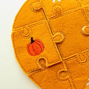 May include: Close-up of a felt puzzle with a pumpkin design. The puzzle pieces are a warm orange colour, with embroidered details and stitching. The pumpkin is orange with a black stem.