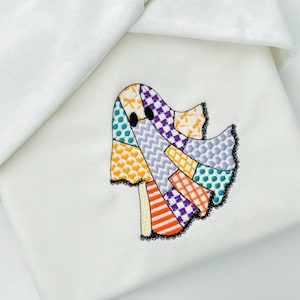 May include: Embroidered ghost design on a white fabric. The ghost is made of colorful patchwork with orange, purple, and teal accents. Black lace trim outlines the ghost. The fabric is folded over.