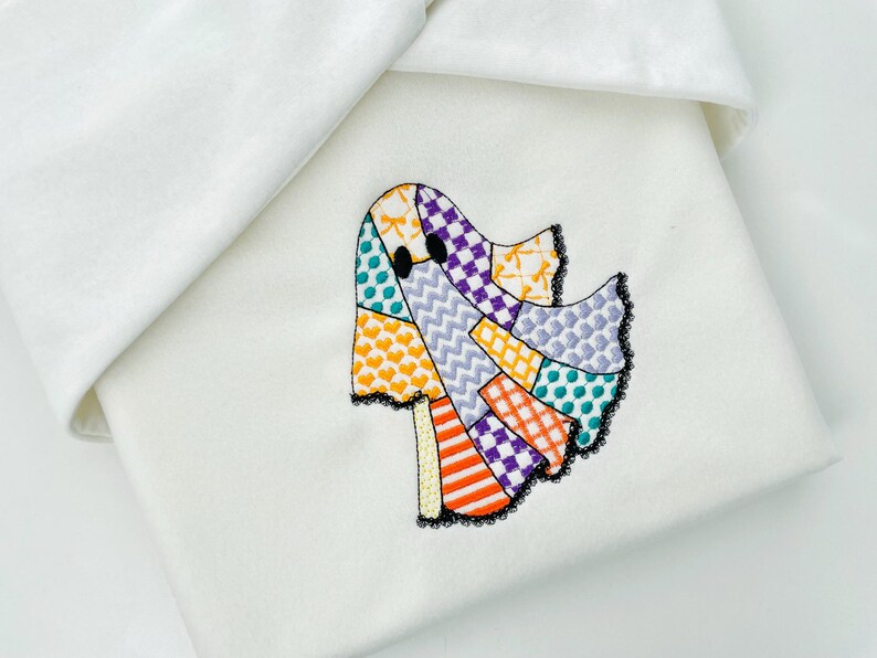 May include: Embroidered ghost design on a white fabric. The ghost is made of colorful patchwork with orange, purple, and teal accents. Black lace trim outlines the ghost. The fabric is folded over.
