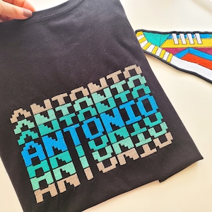 May include: A black t-shirt with a pixelated design of the name "ANTONIO" in shades of blue, green, and beige.