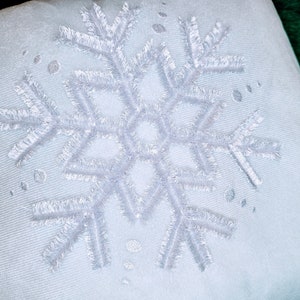 Könnte beinhalten: Ein weißes Kissen mit einem gestickten Schneeflockenmuster.