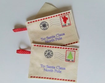 Könnte beinhalten: Zwei Filz-Umschläge mit roten und weißen Tupfenbändern. Die Umschläge sind mit roten und weißen Nähten verziert und tragen die Aufschrift "To: Santa Claus North Pole" mit einem Stempel, auf dem "North Pole" und ein Weihnachtsmotiv abgebildet sind.