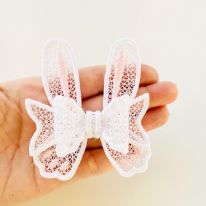 May include: White lace bunny ears with pink accents. The ears are attached to a white lace bow.