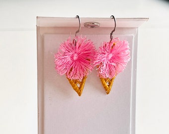 May include: A pair of pink and gold ice cream cone earrings. The earrings are made of yarn and have a fluffy pink top and a gold cone shape.