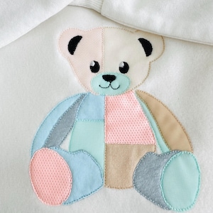 May include: A white sweatshirt featuring a colorful patchwork teddy bear design. The bear has a light pink, blue, beige, and gray color scheme. The bear's eyes and ears are black. The sweatshirt has a white hood.