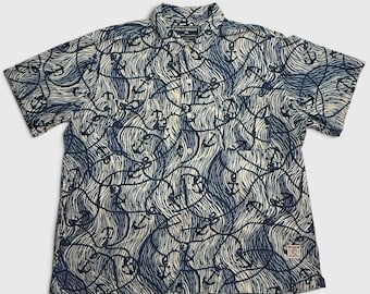 Puede incluir: Una camisa de manga corta con botones en color crema con un patrón azul marino oscuro de líneas onduladas y anclas. La camisa cuenta con cuello, una tapeta de botones frontal y una pequeña etiqueta rectangular de Polo Sport Ralph Lauren.