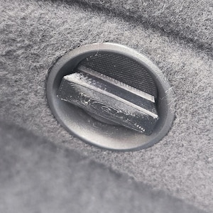 May include: Close-up of a gray, textured fabric with a circular, metallic latch. The latch has a rectangular design and is set into the fabric. The fabric appears to be a soft material.