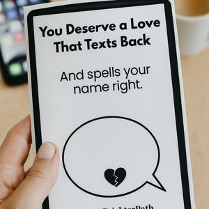 Könnte beinhalten: Eine weiße Karte mit schwarzem Rand, gehalten von einer Hand, zeigt den Text "You Deserve a Love That Texts Back And spells your name right." Eine Sprechblase mit einem gebrochenen Herz-Grafik befindet sich darunter. Die Karte liegt auf einer hellen Holzoberfläche.