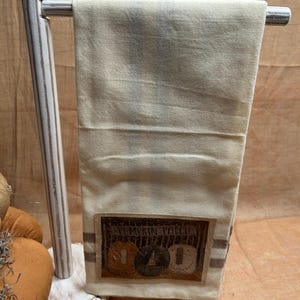 May include: A cream-colored kitchen towel hanging on a white wooden stand. The towel features a rectangular patch at the bottom with the text PUMPKIN PATCH and an illustration of three pumpkins in orange, black, and tan, accented by two horizontal brown stripes.