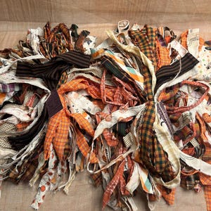 May include: A collection of fabric strips in various autumn-themed patterns including orange, brown, and black plaid, gingham, and pumpkin prints. The strips are frayed at the edges and bundled together in two loose clusters with a rustic aesthetic.