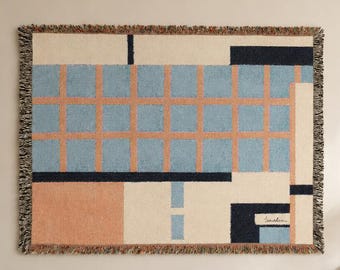 May include: A rectangular woven wall tapestry featuring a geometric grid pattern in light blue, peach, and dark navy blue on a cream background, with a fringed edge and a small signature in the bottom right corner, displayed as a piece of wall art.