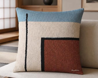 May include: A square throw pillow featuring a textured fabric with a geometric pattern. The design includes a light blue top section, a large beige central area, and a deep red block on the right, all separated by thin black lines.