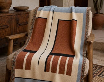May include: A woven throw blanket featuring a geometric pattern in rust orange, cream, and light blue with black linear accents. The blanket has a fringed edge and the word tinatini is embroidered in the bottom right corner of the fabric.