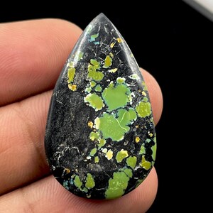 Op de afbeelding: Een druppelvormige cabochon edelsteen met een diepzwarte matrixbasis en levendige limoengroene en geelgroene insluitingen verspreid over het oppervlak. De steen heeft een gladde, gepolijste afwerking met een duidelijk puntige bovenkant.