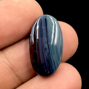 Könnte beinhalten: Ein oval geformter Edelstein mit polierter Oberfläche. Der Stein weist eine Mischung aus dunkelblauen, marineblauen und kastanienbraunen Farbtönen mit einem längs verlaufenden Streifenmuster auf. Der Edelstein wird zwischen Fingern vor schwarzem Hintergrund gehalten.