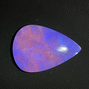Könnte beinhalten: Ein tropfenförmiger Edelstein mit einem faszinierenden Farbspiel. Der Stein weist eine Mischung aus rosa und violetten Farbtönen mit blauen Reflexen auf. Die polierte Oberfläche reflektiert das Licht und erzeugt eine fesselnde Darstellung. Der Edelstein ist vor dunklem Hintergrund.