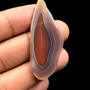 Può includere: Una fetta di pietra d'agata lucidata con una forma naturale e organica. La pietra presenta bande concentriche di colore, tra cui sfumature di arancione, rosso, bianco e grigio. L'agata è tenuta su uno sfondo scuro, evidenziando i suoi intricati motivi e colori.