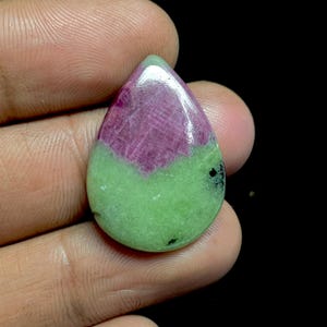 Puede incluir: Una gema en forma de lágrima con un contraste de color vibrante. La mitad superior es de un magenta intenso, mientras que la mitad inferior es verde claro. La piedra se sostiene entre los dedos sobre un fondo negro.