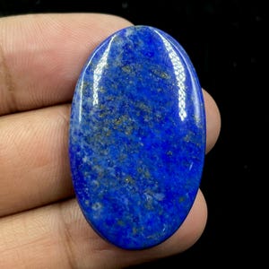 May include: A polished, oval-shaped lapis lazuli gemstone. The deep blue stone has gold flecks and a glossy finish. The gemstone is held in a hand against a black background. This stone is often used in jewelry.