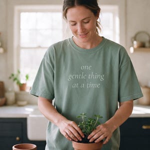 May include: A sage green short-sleeve t-shirt featuring the text 'one gentle thing at a time' printed in a light color on the chest. The shirt has a crew neckline and a relaxed fit, constructed from a solid-colored cotton fabric.