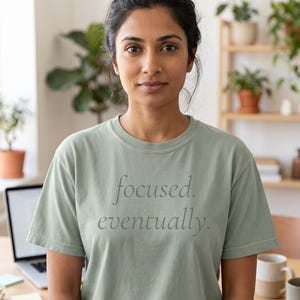 May include: A sage green short-sleeved crew neck t-shirt featuring the text "focused. eventually." printed in a dark grey serif font across the chest. The shirt is made of a solid-coloured fabric with a standard fit and a ribbed neckline.