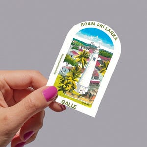 May include: A white sticker featuring a watercolour illustration of a lighthouse in Galle, Sri Lanka. The sticker has the text "ROAM SRI LANKA" at the top and "GALLE" at the bottom. Palm trees and buildings are also visible.