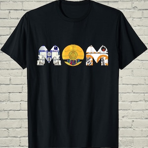May include: Black t-shirt featuring the word "MOM" in a Star Wars-themed design. The letters are stylized to resemble R2-D2, C-3PO, and BB-8. The shirt is made of a soft, comfortable material.