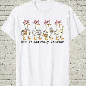 May include: White t-shirt featuring a cartoon design of four geese with pink bows, each carrying different items. The geese are walking and the text "Off To Actively Monitor" is printed below the geese.
