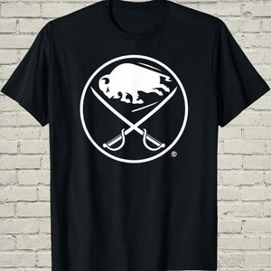 May include: Black t-shirt featuring a white circular logo with a buffalo and crossed sabers. The buffalo is centered within the circle, and the sabers are positioned diagonally. The design is a sports team logo.