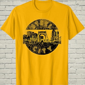 May include: Mustard yellow t-shirt featuring a distressed black graphic of a city skyline and bridge within a circle. The text "STEEL CITY" is arched above and below the graphic. The shirt has a crew neck and short sleeves.