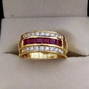 May include: A gold band ring featuring a central row of five square-cut deep red rubies flanked by two rows of small, round, clear white stones. The metal has a polished yellow finish, and the design is symmetrical with a wide, structured profile.