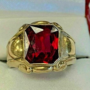 May include: A gold ring featuring a large, rectangular, faceted deep red gemstone set in the center. The band has a smooth, polished gold finish with decorative, rounded metal accents surrounding the base of the central stone setting.