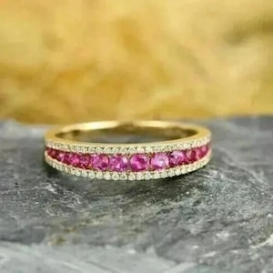 May include: A gold band ring featuring a central row of round, vibrant pink gemstones flanked by two thinner rows of small, clear white stones. The metal has a polished finish, and the design showcases a symmetrical arrangement of stones.