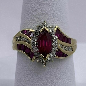 May include: A gold ring featuring a marquise-cut ruby center stone surrounded by a halo of diamonds. The ring has additional ruby accents and diamond details on the band, creating a luxurious and elegant design.
