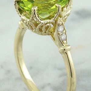 May include: A gold ring featuring a large, oval-cut, light green gemstone. The gemstone is set in a detailed, ornate gold setting with small accent diamonds. The ring's band is a simple, polished gold.