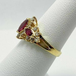 May include: A gold ring featuring two marquise-cut red gemstones and several small, round, clear gemstones. The ring is set on a white background, showcasing its intricate design and sparkling details.