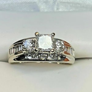 May include: A white gold engagement ring with a large square-cut center stone, flanked by two smaller round diamonds and baguette-cut diamonds on the band. The ring is set in a white setting.