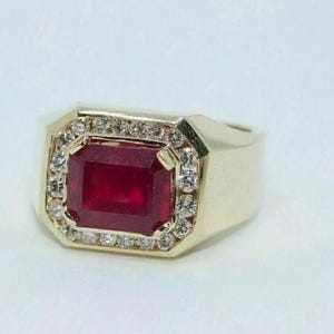 May include: A gold ring featuring a large, rectangular, deep red ruby set in the center. The ruby is surrounded by a halo of small, round, clear white diamonds. The band of the ring is wide and made of polished gold, creating a bold, solid appearance.