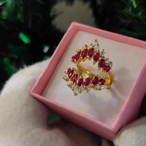 May include: A gold-toned ring with a split shank design, adorned with alternating ruby-red and clear gemstones. The ring is presented in a pink jewelry box, showcasing its intricate details and vibrant colors.