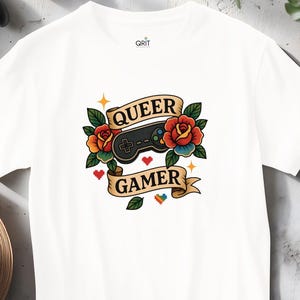 May include: White t-shirt with a colorful graphic featuring a black game controller, roses, and the words "QUEER GAMER" on a banner. A black game controller and a straw hat are visible in the background.