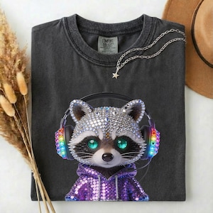 May include: Dark gray t-shirt featuring a rhinestone-covered raccoon wearing headphones with rainbow accents. The raccoon is wearing a purple sequined hoodie. A silver necklace with a star pendant is on the shirt.