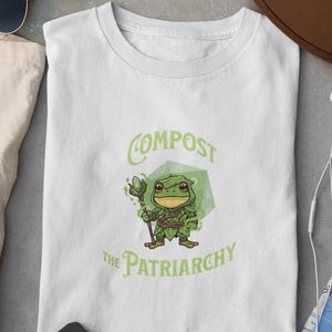 May include: White t-shirt with a green cartoon frog holding a staff, set against a green geometric shape. The shirt features the text "COMPOST THE PATRIARCHY" in green, with a whimsical font.