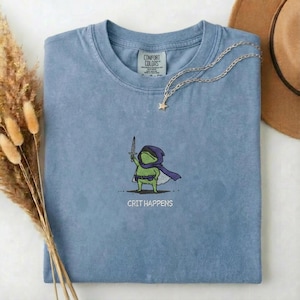May include: A blue crew neck t-shirt featuring an embroidered graphic of a green frog wearing a purple cloak and holding a sword. Below the frog, the text reads CRIT HAPPENS. The shirt has a Comfort Colors label at the inner neckline.