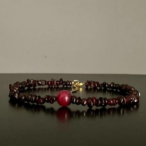 May include: A dark red beaded bracelet with a gold clasp and a large, round, red bead in the centre. The bracelet is made of small, irregularly shaped beads. The background is a dark surface and a light grey wall.