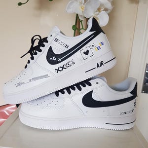 May include: A pair of white sneakers with black laces and black Nike swoosh logos. The upper features black graphic designs including a heart, the text AIR, and the phrase HANDLE WITH CARE. The shoes have a thick white rubber sole and black heel tabs.