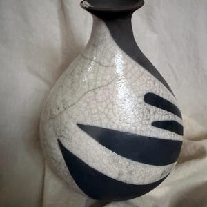 May include: A ceramic vase with a bulbous body and a narrow neck. The vase is primarily white with a crackle glaze, and features a black rim and black, curved, abstract shapes. The vase is approximately 8 inches tall.