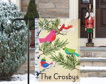 May include: A decorative garden flag featuring colorful birds perched on pine branches. The flag has a cream background and includes the text "The Crosbys" at the bottom. The birds are in shades of pink, blue, green, and white.