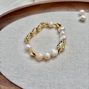 May include: A gold-toned bracelet featuring a combination of chain links and organic-shaped pearls. The bracelet has a mix of textures, with the smooth pearls contrasting the textured metal. The bracelet is displayed on a neutral linen surface.